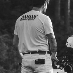 Model wearing Motorcycle Desert Race white t-shirt with a Bike Shed Moto logo, standing on a motorcycle in a forest.