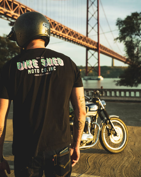 Model wearing a 'Bike Shed Moto Co.' graphic logo t-shirt with a motorcycle and bridge in the background.