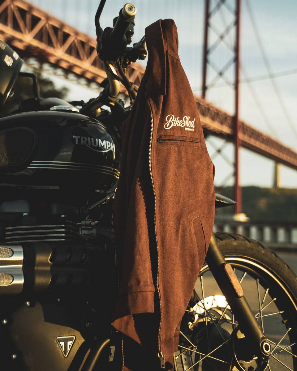 The Bike Shed Moto Co Twill brown jacket with a Bike Shed brand logo hanging from motorcycle handlebars.