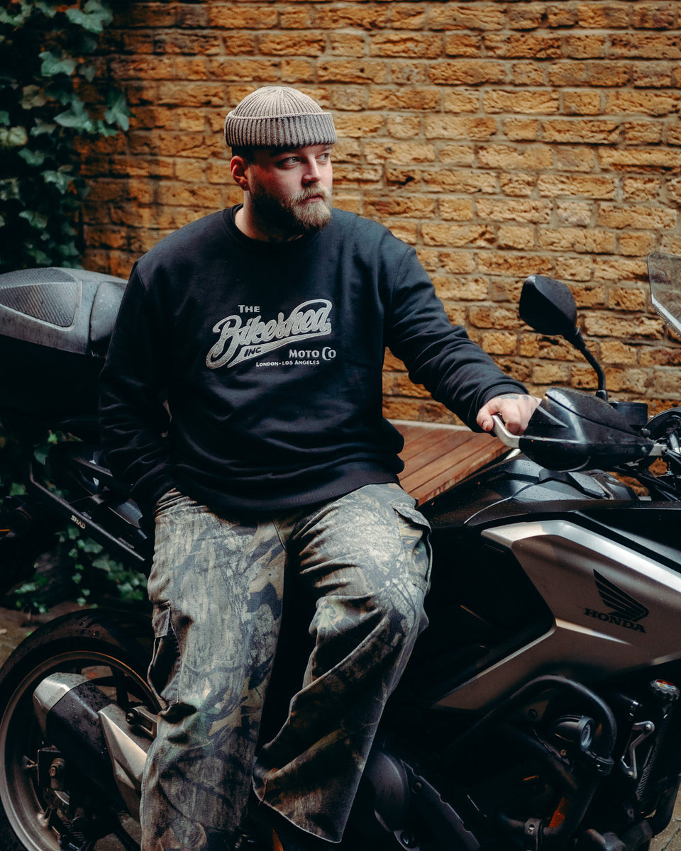 Model wearing Bike Shed Moto Co Black Applique Sweatshirt next to a motorcycle on a brick driveway