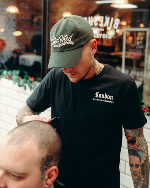 Model wearing black Bike Shed Moto Co Olde London Logo T Shirt while cutting hair in a barbershop