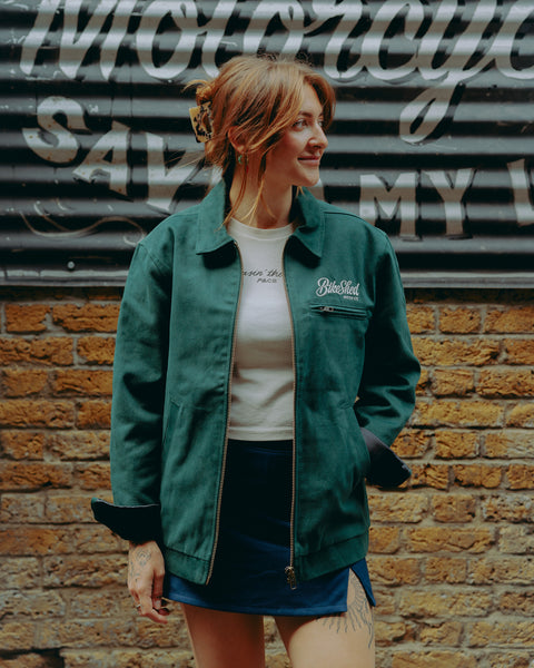 Model wearing green motorcycle jacket in front of a brick wall with graffiti.