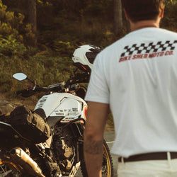 Model wearing Motorcycle Desert Race white t-shirt with a Bike Shed Moto logo, standing next to  a motorcycle in a forest.