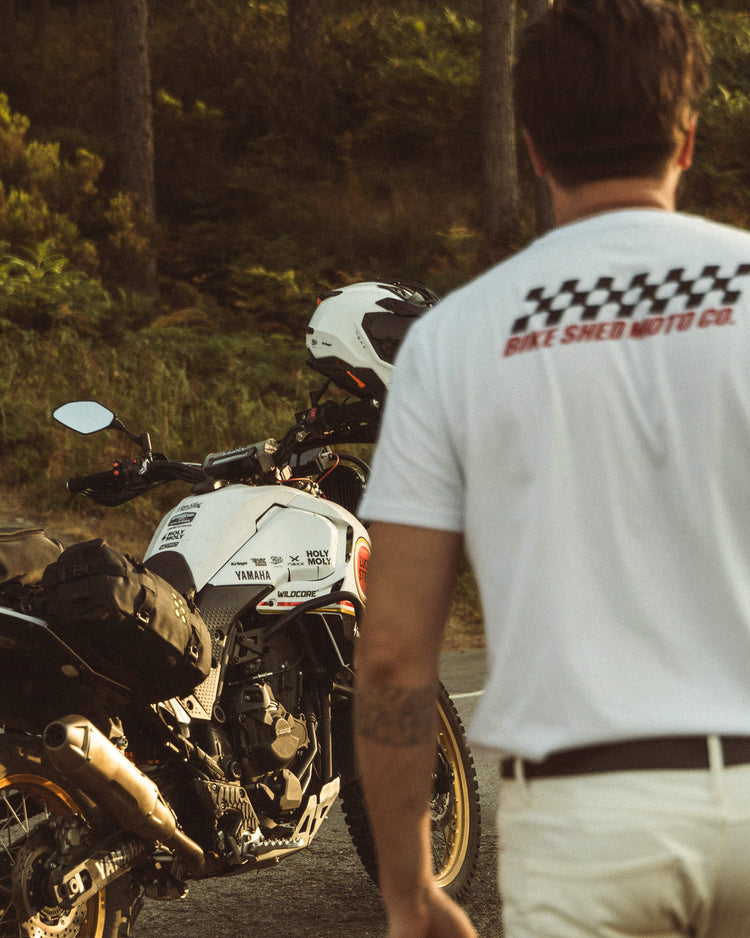 Model wearing Motorcycle Desert Race white t-shirt with a Bike Shed Moto logo, standing next to  a motorcycle in a forest.