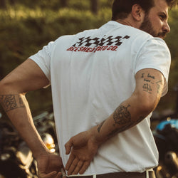 Model wearing Motorcycle Desert Race white t-shirt with a Bike Shed Moto logo, standing in a forest.