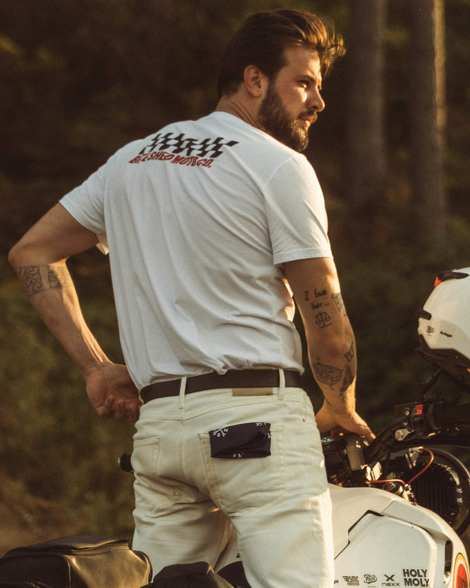 Model wearing Motorcycle Desert Race white t-shirt with a Bike Shed Moto logo, standing on a motorcycle in a forest.