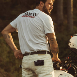 Model wearing Motorcycle Desert Race white t-shirt with a Bike Shed Moto logo, standing on a motorcycle in a forest.