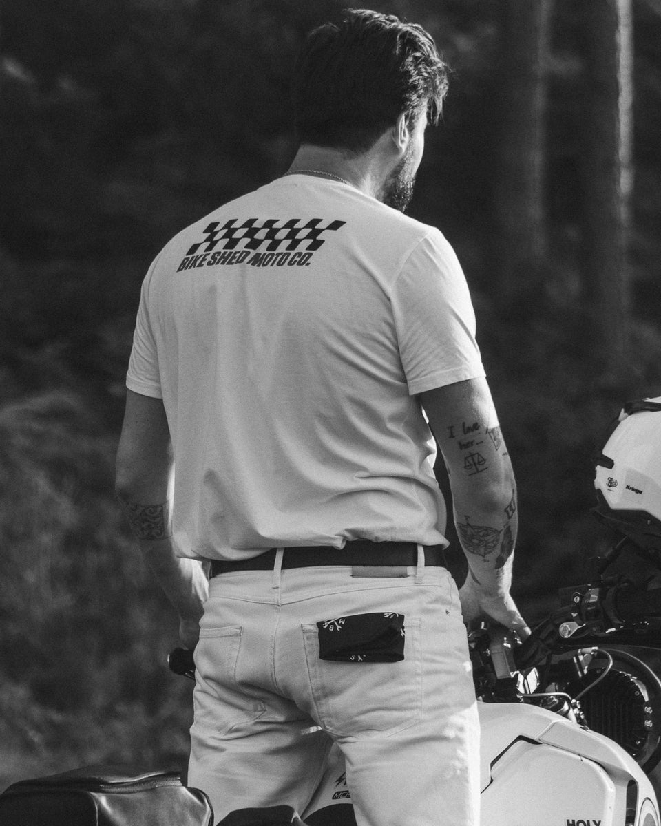 Model wearing Motorcycle Desert Race white t-shirt with a Bike Shed Moto logo, standing on a motorcycle in a forest.